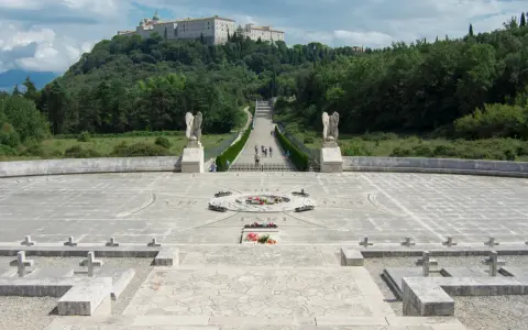 Cmentarz żołnierzy na wzgórzu, z widocznym klasztorem na szczycie. Droga prowadzi do niego przez monumentalny plac z krzyżami i pomnikiem. To miejsce pamięci o bitwie pod **Cassino - trzeci / rzeka**.