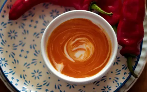 Crema di peperoni vellutata, con un vortice di panna, accanto a peperoni rossi freschi su un piatto decorato.