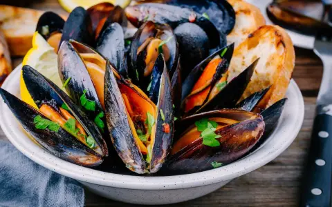 Mejillones en salsa, servidos en un cuenco blanco con perejil fresco, rodajas de limón y pan tostado.