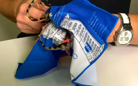 Brille mit buntem Rahmen wird mit einem blauen Tuch gereinigt. So lässt sich Haarspray von Brille entfernen.