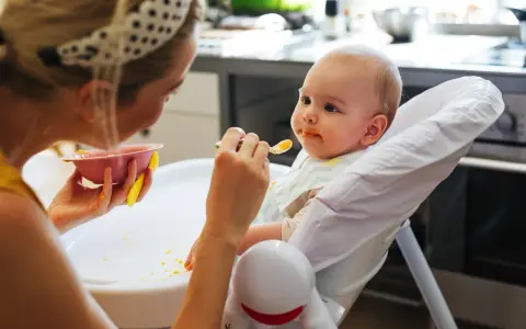 Mama karmi 7 miesięczne dziecko puree z marchewki. Maluch ma pomarańczowe ślady na buzi i śliniaczku.