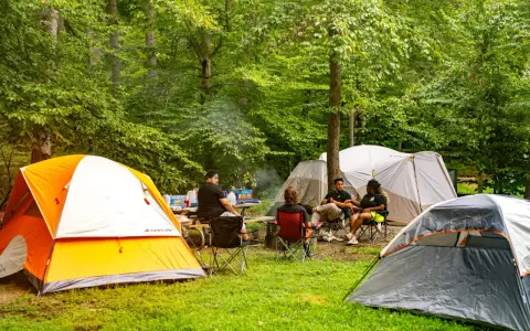 Grupa przyjaciół odpoczywa na polu namiotowym Mazury. Wokół rozstawione są namioty, a na stole piknikowym stoją napoje i przekąski.