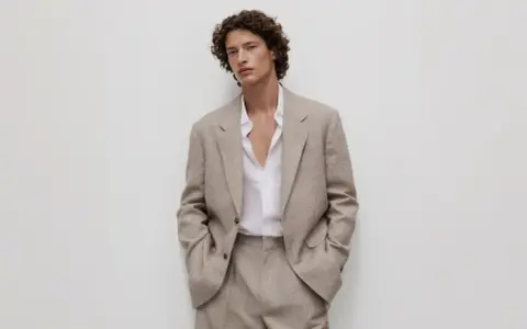 Uomo con capelli ricci indossa un completo beige, incarnando l'eleganza discreta dell'old money significato.