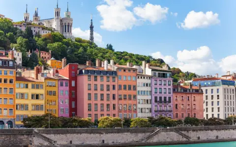 Colorful buildings line the river in Lyon, France, with a basilica on the hill above. A perfect scene for Lyon travel.