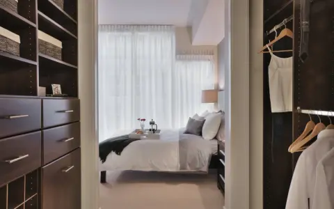 Dormitorio con vestidor integrado. Un espacio organizado con estanterías oscuras y cajones, que da paso a una cama acogedora y un armario con ropa colgada.