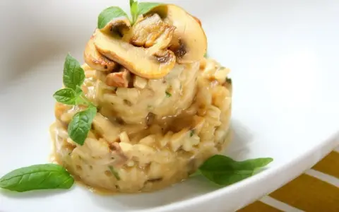Kremowe risotto z grzybami, udekorowane listkami bazylii i oregano, prezentuje się apetycznie na białym talerzu.