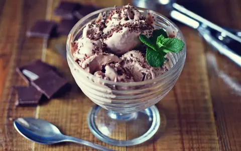Pyszne lody nuii w pucharku, posypane czekoladą i udekorowane miętą. Obok leżą kawałki czekolady i łyżeczka.