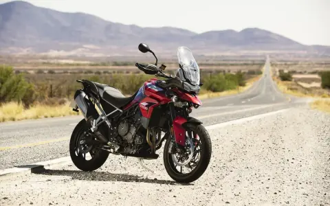 Czerwony motocykl Triumph Tiger 900 GT Pro zaparkowany na poboczu pustej drogi w krajobrazie z górami w tle.