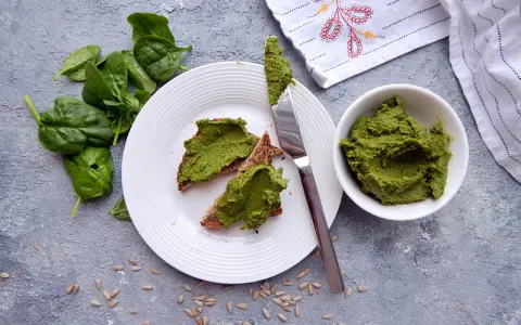 Pesto ze szpinaku do słoików na chlebie z pestkami słonecznika. Świeży szpinak obok talerza.