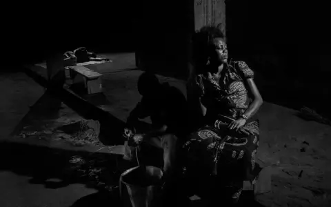 A woman in a patterned dress sits in shadow, eyes closed, while a man works with a bucket in the foreground. Alex Majoli captures a moment of quiet resilience.