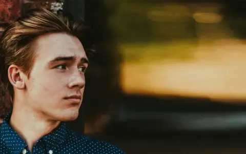 Junger Mann mit stylischen herren frisuren mittellang, lehnt an einer Ziegelwand.