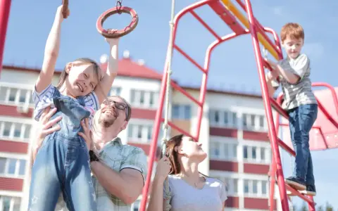Radosna rodzina na placu zabaw. Ojciec podtrzymuje córkę na kółkach gimnastycznych, syn wspina się po drabince. Zabawy na placu zabaw cele to radość i wspólne chwile.