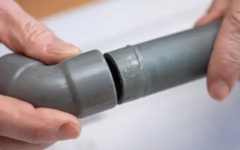 Mains de personne assemblant deux tuyaux gris en **pvc renforcé**, l'un avec un coude, prêts pour une installation solide.