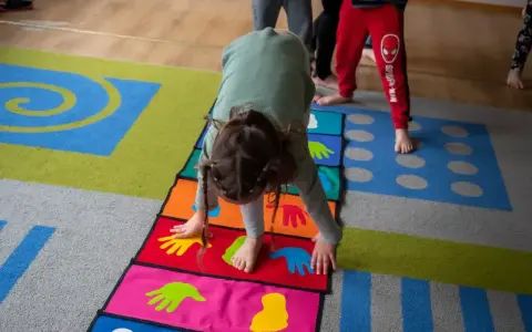 Dziecko wykonuje ćwiczenia na kolorowej macie z symbolami dłoni i stóp. Poduszka sensoryczna wspiera rozwój motoryczny.