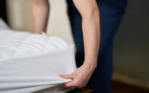 Dłoń naciąga biały ochraniacz na materac, ukazując jego pikowany szew materacowy.