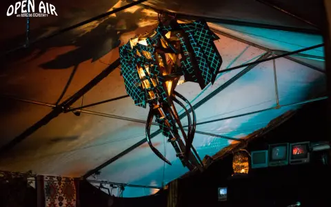 Abstrakte Skulptur mit blauem Gittermuster unter einem Zeltdach, beleuchtet von warmem und kühlem Licht.
