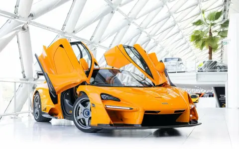 Ein leuchtend oranger McLaren Senna LM mit geöffneten Flügeltüren steht in einem modernen Gebäude mit Glasdach.
