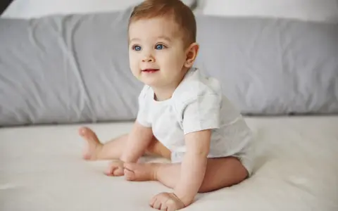 Niebieskooki niemowlak w jasnym body siedzi na łóżku, patrząc w bok.