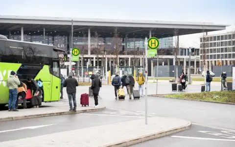 Ludzie z bagażami wsiadają do zielonego autokaru przed nowoczesnym budynkiem, przypominającym lotniska w Berlinie.