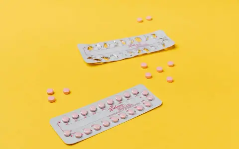 Dos blísteres de pastillas anticonceptivas y varias pastillas sueltas, rosas, sobre fondo amarillo. Ayudan a regular la regla.