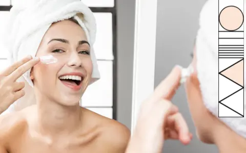 Donna sorridente con asciugamano in testa applica crema sul viso, parte della sua routine skincare.