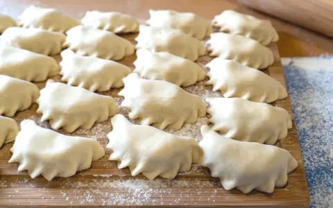 Surowe pierogi ułożone rzędami na drewnianej desce, posypanej mąką.