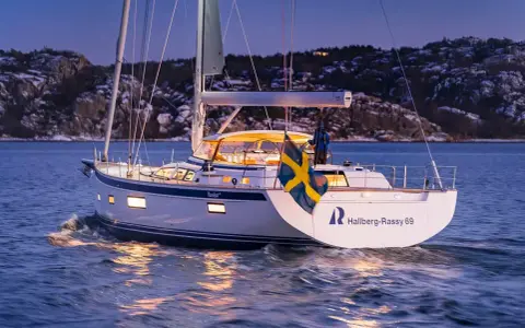 Eine Hallberg-Rassy 69 gleitet bei Dämmerung über das Wasser, die schwedische Flagge weht im Wind.