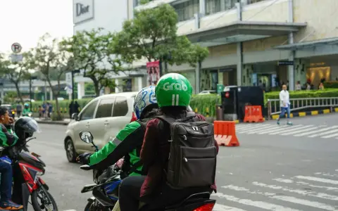 Zwei Personen auf einem Roller, der Fahrer trägt einen grünen Helm mit dem "Grab" Logo. Sie sind unterwegs in Thailand, um mit der Grab App Kunden zu transportieren.