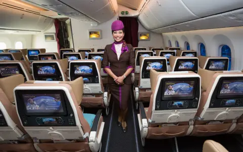 Uśmiechnięta stewardessa Etihad Airways w brązowym mundurku stoi w rzędzie foteli. Ekraniki pokazują astronautę. Dobre etihad airways opinie.