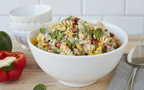 Ein bunter Nudelsalat ohne Mayonnaise mit Spiralnudeln, Erbsen, Mais, Paprika und frischem Basilikum in einer weißen Schüssel.