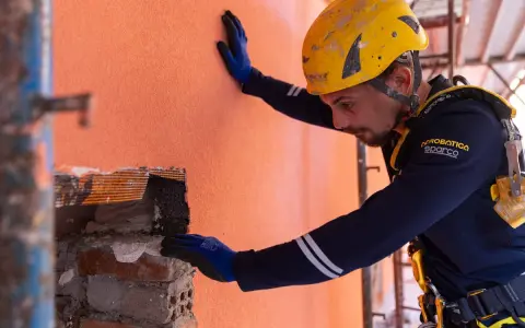 Un ouvrier en casque jaune effectue des travaux d'isolation extérieur, appliquant un enduit sur un mur orange.
