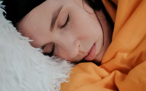 Viso di donna con gocce di sudore sulla fronte, avvolta in una coperta arancione e piumino bianco. Sembra che stia per sudare freddo.