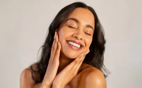 Une femme au sourire radieux caresse son visage, révélant une peau glowy éclatante de santé.