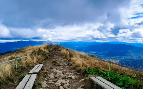 Kamienista ścieżka prowadzi przez trawiaste wzgórza, oferując zapierające dech w piersiach **Bieszczady widoki**. Na pierwszym planie drewniane ławki, w oddali błękitne pasma górskie pod burzowym niebem.