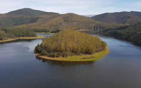 El meandro del Melero, una isla verde rodeada de agua serena, se abre paso entre colinas boscosas bajo un cielo azul.