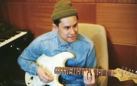 Frank Dukes w zielonej czapce i dżinsowej koszuli gra na gitarze Fender Stratocaster w studiu.