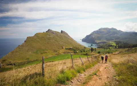 Two hikers with backpacks walk on a dirt path through a grassy landscape with mountains and the sea in the background.