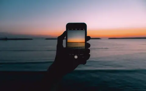 Dłoń trzyma smartfon, na ekranie którego widać piękny zachód słońca nad morzem. Idealny kadr, by nauczyć się, jak fotografować zachód słońca.