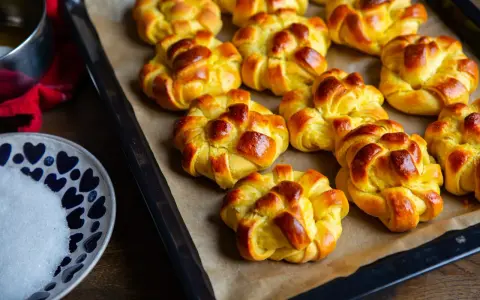 Nybakade lussebullar med kesella, gyllene och redo att avnjutas. En skål med socker och en röd duk anas i bakgrunden.
