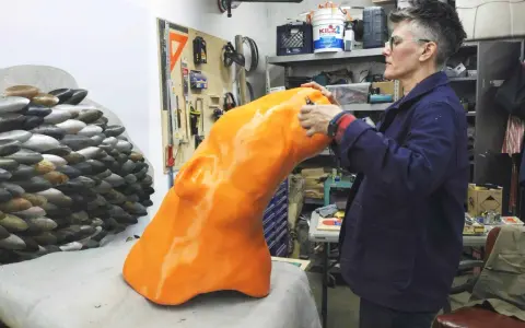 Artist applies finishing touches to a large, orange, 3D printed object, part of a project creating large objects.