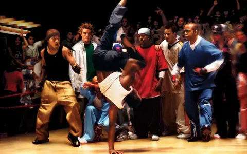 Energetyczna scena z filmów o tańcu. Breakdancer wykonuje akrobację, otoczony przez widzów i innych tancerzy.