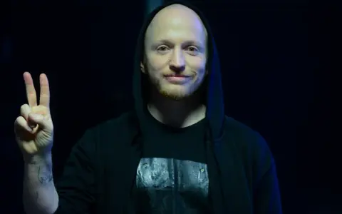 Wojtek Urbański w czarnej bluzie z kapturem pokazuje znak pokoju.