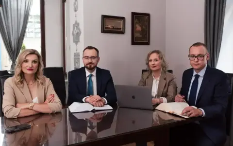 Cztery osoby w strojach biznesowych siedzą przy stole, przed nimi dokumenty i laptop.