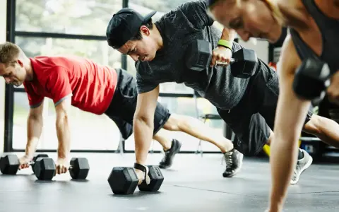 Drei Personen machen ein intensives Crossfit Training mit Kurzhanteln. Sie sind in einer Plank-Position und ziehen abwechselnd eine Hantel hoch.