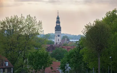 Wieża zegarowa góruje nad zieloną górą, skąd rozciąga się widok na rynek i dachy domów.