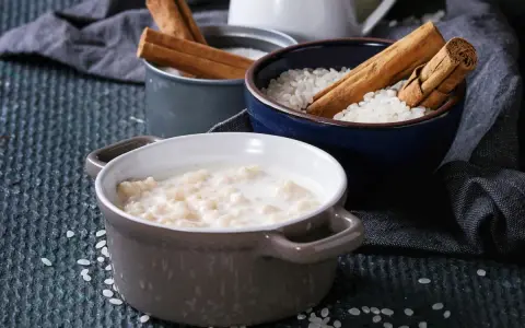 Gotowy do podania ryż na mleku w ceramicznej miseczce, obok miseczki z ryżem i laskami cynamonu.