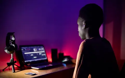Un jeune homme, casque sur les oreilles, est assis devant un ordinateur portable, prêt à créer un son unique dans une ambiance tamisée aux lumières violettes et rouges.