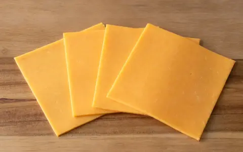 Vier Scheiben gelber Cheddar-Käse, laktosefrei, liegen auf einem Holzbrett.