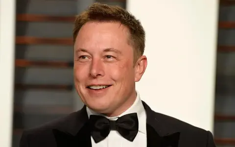 Uśmiechnięty Elon Musk w smokingu z muszką. Wiek nie jest widoczny, ale jego twarz wyraża pewność siebie.