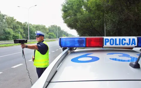 Policjant z radarem na drodze, obok radiowozu z napisem POLICJA.